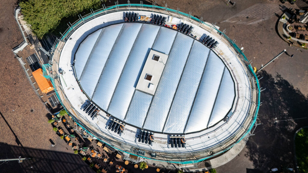 Bovenaanzicht van het ovale JinSo-gebouw met de centrale overkapping, witte dakbedekking rondom en steigers langs de gevel.